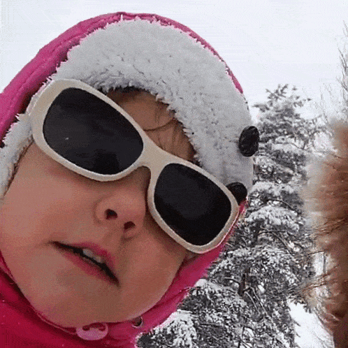 a little girl wearing sunglasses and a pink hat is standing in the snow