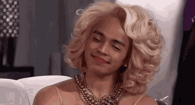 a man wearing a blonde wig and a pearl necklace is smiling .