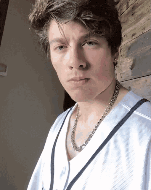 a young man wearing a white shirt and a chain around his neck looks at the camera