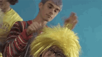 a man is brushing another man 's hair while wearing a yellow wig and glasses .