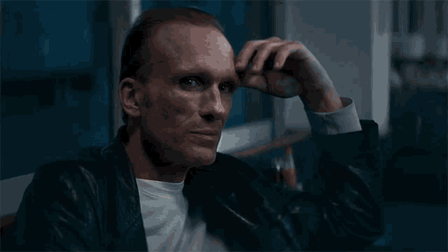 a man in a leather jacket is sitting at a table with his hand on his forehead