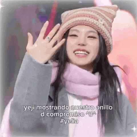 a woman wearing a hat and scarf is smiling and waving while wearing a ring on her finger .