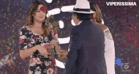 a woman in a floral dress is holding a rose and shaking hands with a man in a suit .