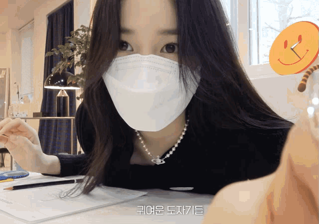 a woman wearing a face mask is sitting at a desk with a smiley face on the wall behind her