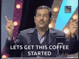 a man in a suit and glasses is standing in front of a microphone and saying let 's get this coffee started .