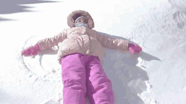 a little girl in a pink jacket and pink pants is laying in the snow