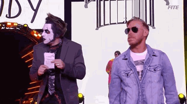 two men are standing next to each other on a stage . one of the men is wearing a mask and sunglasses .