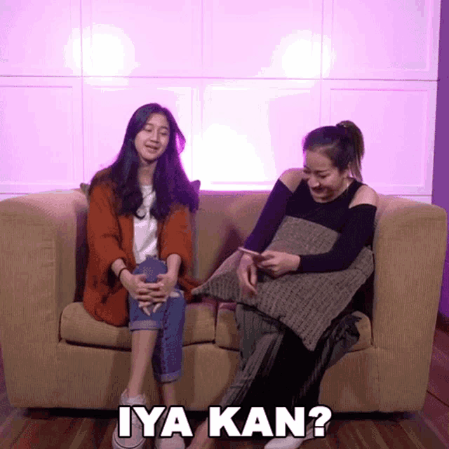 two women are sitting on a couch and one of them is using a cell phone and the other is asking " iya kan "