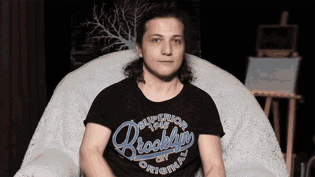 a woman wearing a black shirt that says brooklyn city original sits in a chair