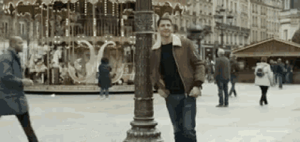 a man in a red jacket is standing next to a street light in front of a merry go round .