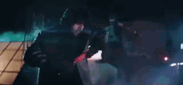 a man in a hooded jacket is holding a gun in a dark room and says chal hat .