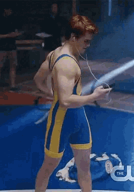 a man in a blue and yellow wrestling uniform is standing on a blue mat looking at his phone .