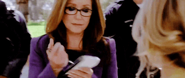 a woman in a purple jacket and glasses is holding a piece of paper in her hand .