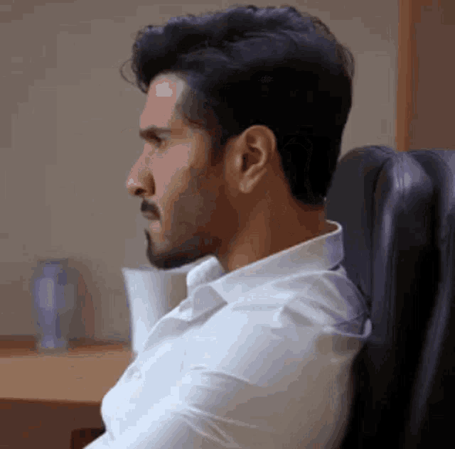 a man with a beard is wearing a white shirt and sitting in a chair