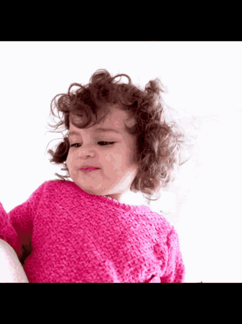 a little girl with curly hair wearing a pink sweater looks at the camera