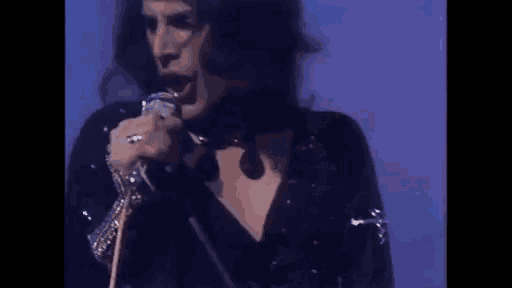 a man with long hair is singing into a microphone while standing in front of a blue wall .