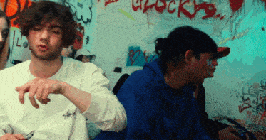 a man wearing a stussy sweatshirt is sitting in front of a wall that has graffiti on it