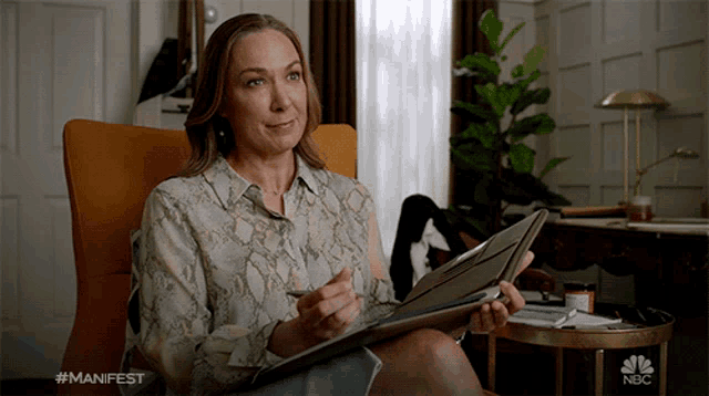 a woman is sitting in a chair holding a folder with the nbc logo visible