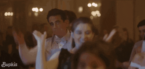 a man in a tuxedo and bow tie is pointing at something in a crowd of people with the word bupkis on the bottom left