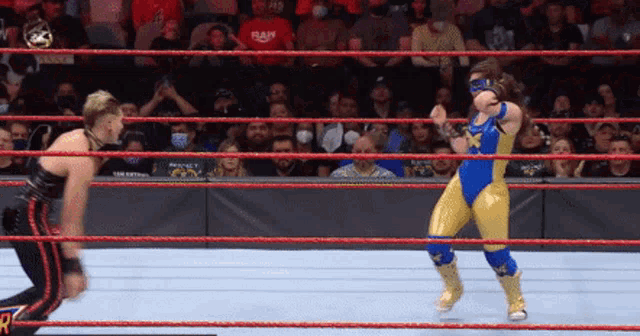 a wrestler in a blue and gold outfit is jumping in the air while another wrestler watches