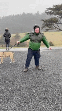 a man in a green jacket is walking a dog on a leash on a gravel road .