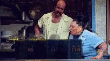 a man and a woman in a diner behind a counter looking at menus