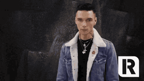 a young man wearing a denim jacket with a fur collar is standing in front of a black wall .