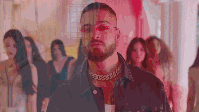 a man wearing sunglasses and a chain around his neck stands in front of a crowd of women