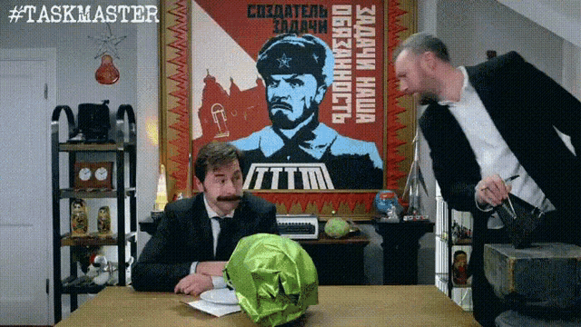 two men are sitting at a table in front of a large poster that says taskmaster