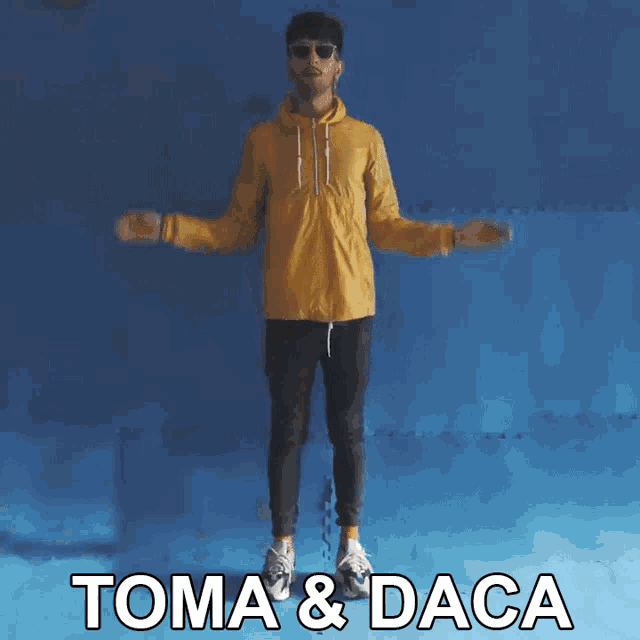 a man in a yellow jacket stands in front of a blue wall with the words toma & daca