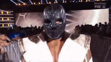 a wrestler wearing a skull mask and a white jacket is walking on stage .