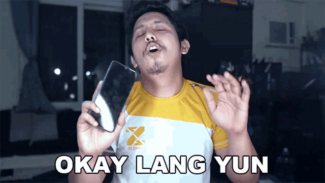 a man in a yellow and white shirt holds a cell phone and says " okay lang yun "