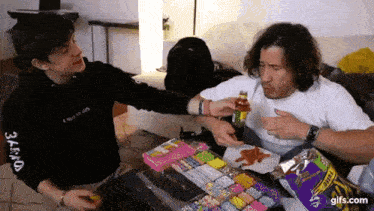 two men are sitting at a table eating candy and one of them is holding a bottle of juice .