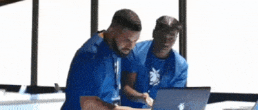 two men in blue shirts are looking at a laptop .