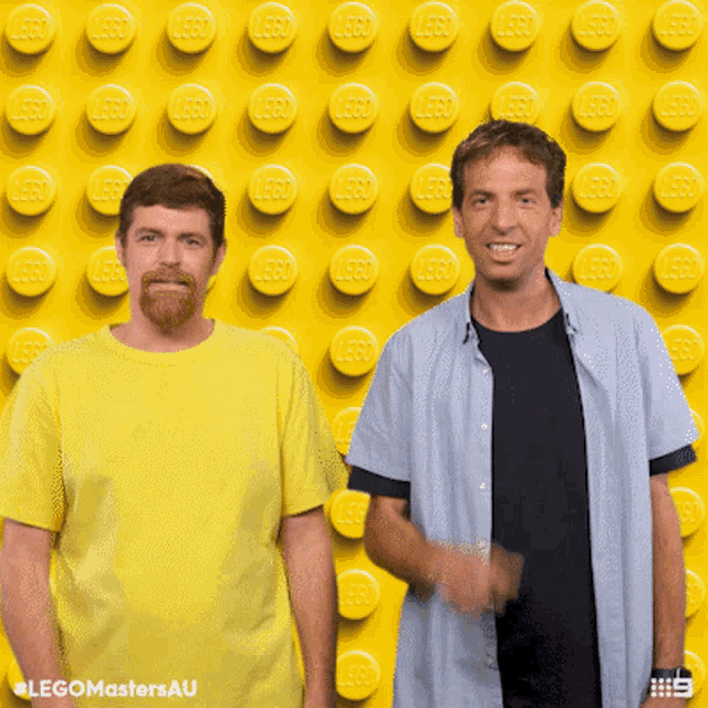 two men standing in front of a wall of yellow lego blocks