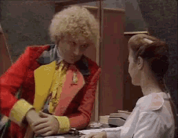 a man in a red and yellow jacket is talking to a woman in a white shirt .