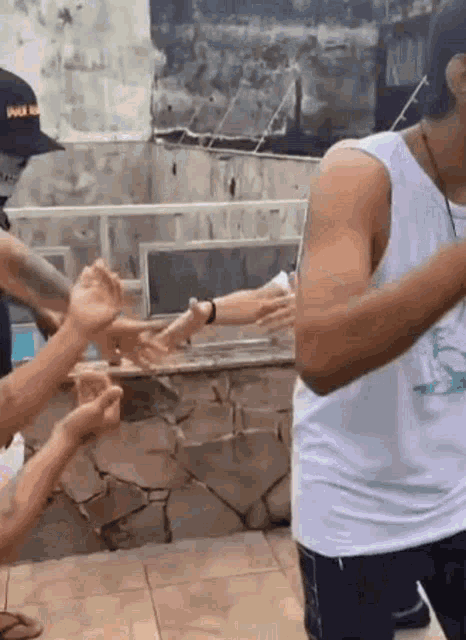 a man in a white tank top is standing next to a group of people reaching out their hands .