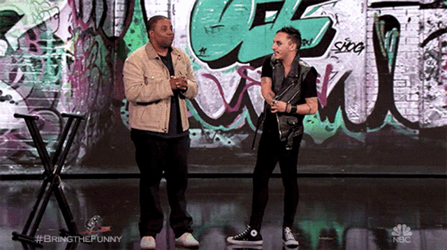 two men stand on a stage in front of a wall that has graffiti on it and the words bring the funny on the bottom
