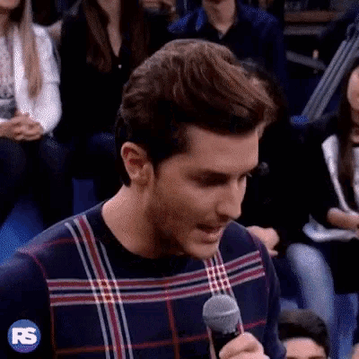 a man wearing a plaid sweater is holding a microphone in front of a crowd .
