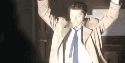a man in a trench coat and tie is standing in a dark room with his arms in the air .