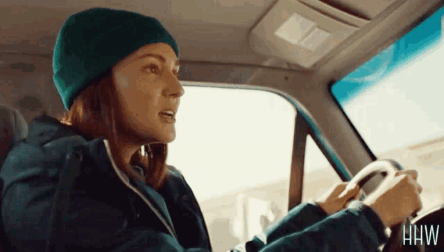 a woman wearing a green beanie is driving a car with hw written on the steering wheel