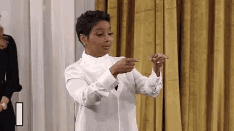 a woman in a white shirt is pointing at something while standing in front of a curtain .