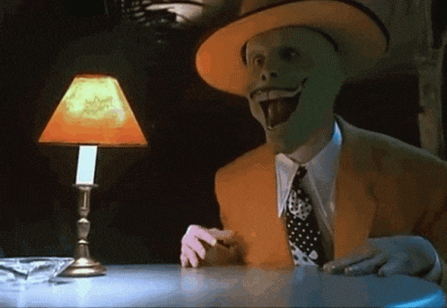 a man with a mask on his face is sitting at a table with a lamp behind him