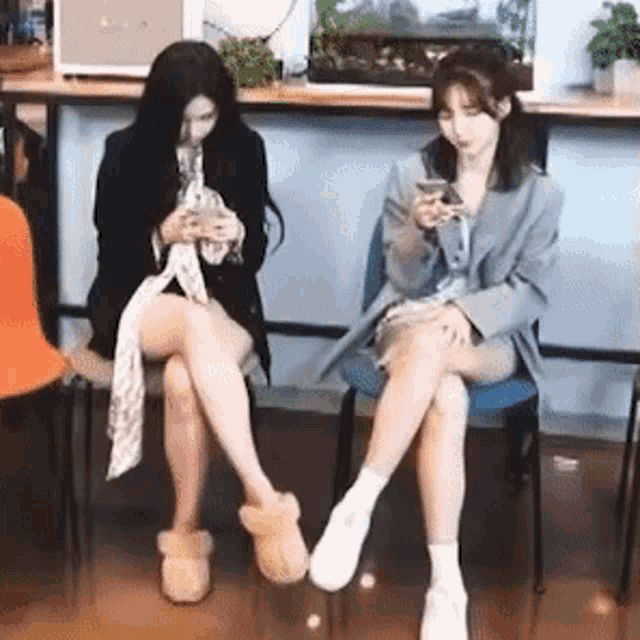 two women are sitting on chairs with their legs crossed looking at their phones .