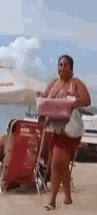 a woman in a bikini is standing on a beach carrying a cooler .