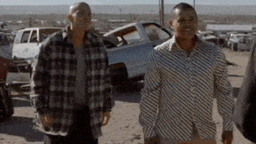 two men in plaid shirts are standing next to each other in a junkyard .