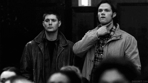 a black and white photo of two men standing next to each other in a crowd . one of the men is pointing at the camera .