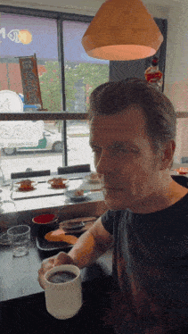 a man sitting at a table with a cup of coffee in front of a window with a sign that says 2sm on it