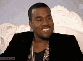 a man in a black jacket and gold chains is smiling while sitting on a bed .