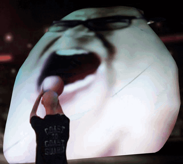 a man wearing a coast guard shirt stands in front of a giant screen